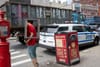 A man holding a black box stands at a crosswalk next to which a police car is parked and a truck is driving by